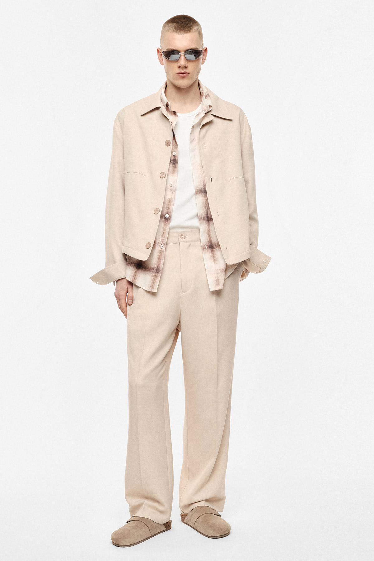Young man in beige relaxed trousers and jacket, checked shirt over white tee, sunglasses and beige slippers in studio