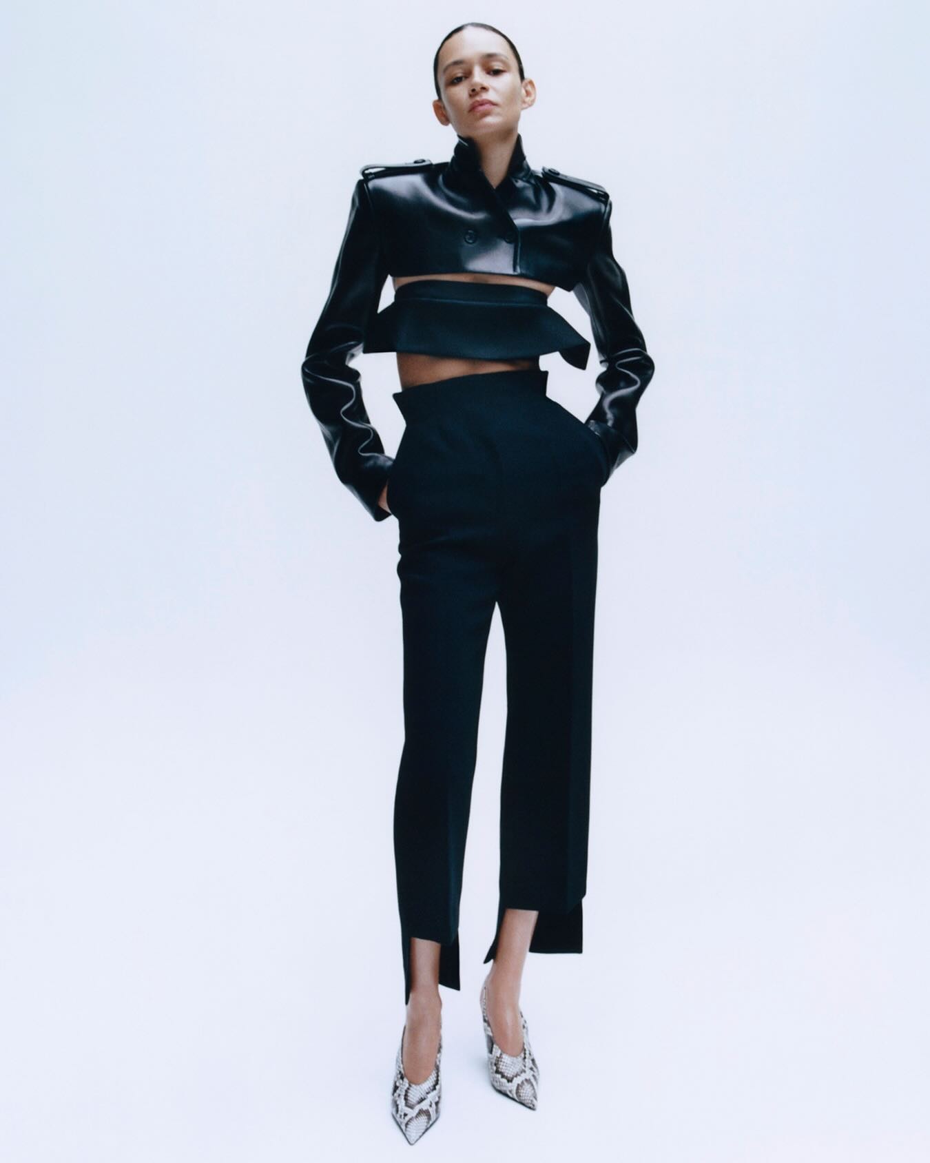 Model in a cropped black leather jacket, high-waisted black trousers with ankle slits and snakeskin heels on a pale background