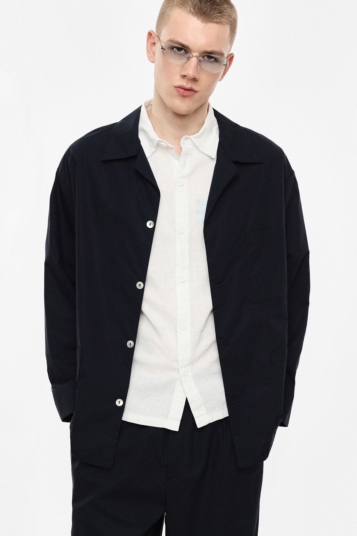 Young male model wearing a dark navy overshirt, white button-up shirt, black trousers and silver-rim glasses against a plain studio background