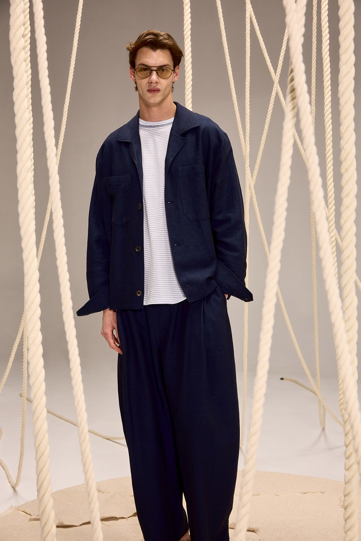 Male model in navy relaxed suit and striped T-shirt with sunglasses standing amid hanging thick ropes on a neutral backdrop