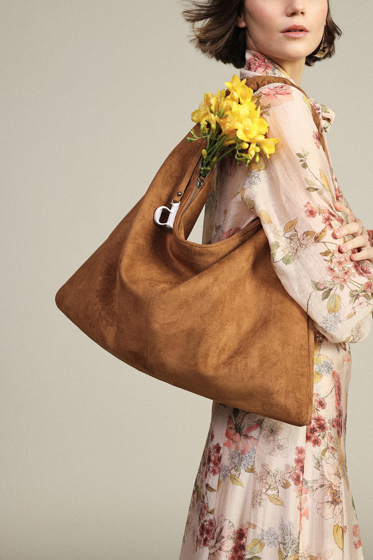 Woman in floral dress carrying tan suede hobo bag with yellow freesias peeking out, fashion accessory