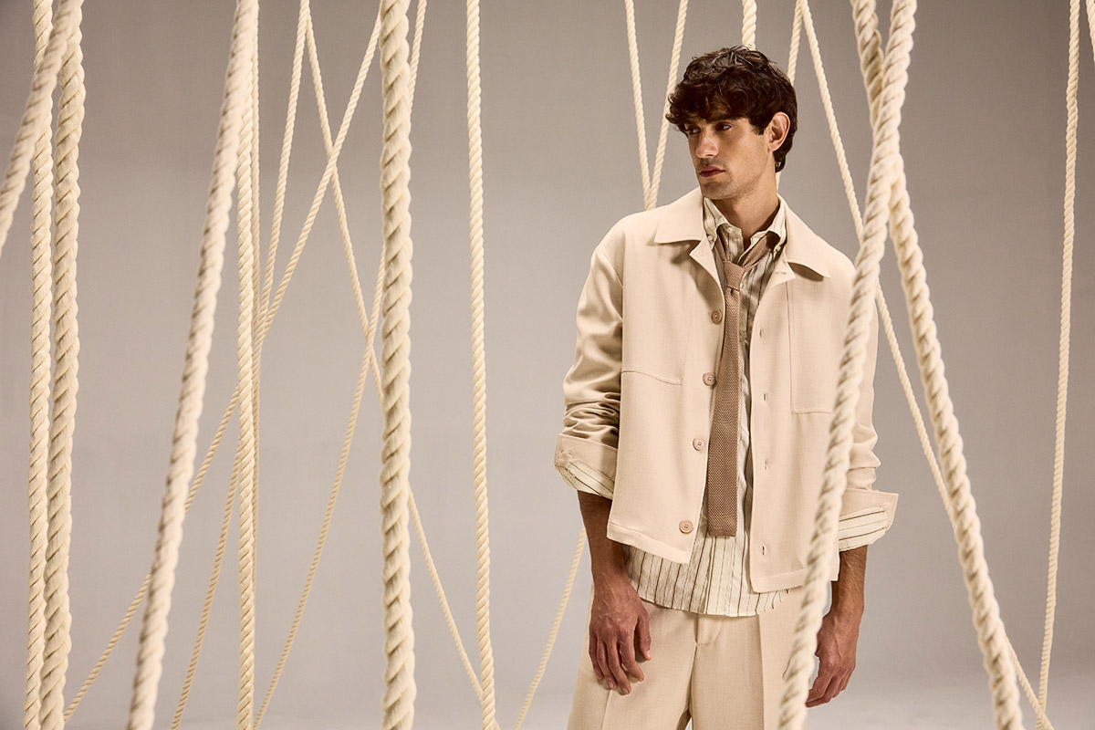 Male model in beige jacket, shirt and trousers standing among hanging thick ropes in a minimalist studio fashion photoshoot