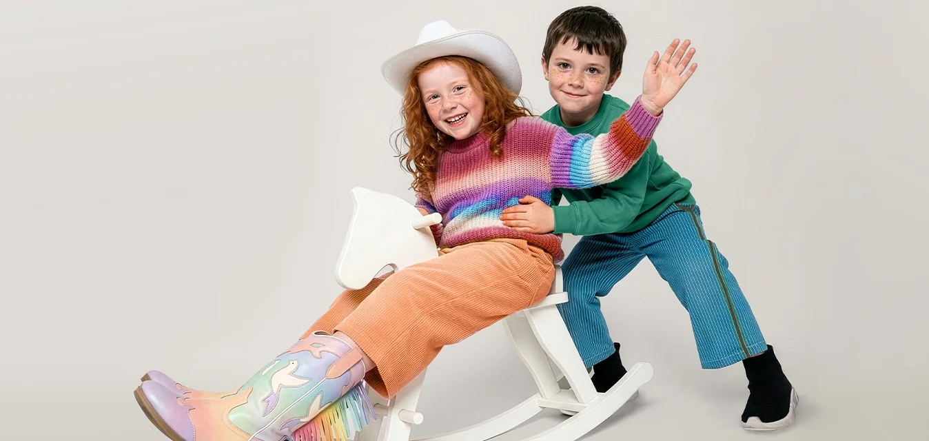 Due bambini giocano con un cavallo a dondolo in legno bianco su uno sfondo neutro; un bambino saluta mentre l'altro spinge.