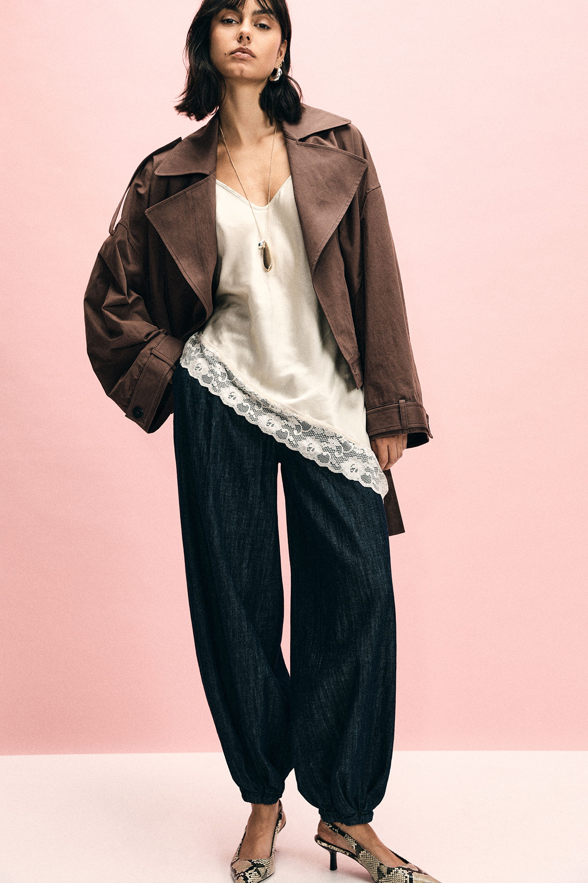 Woman modelling oversized brown coat, silk lace‑trim camisole, loose dark trousers and slingback heels against a pale pink background