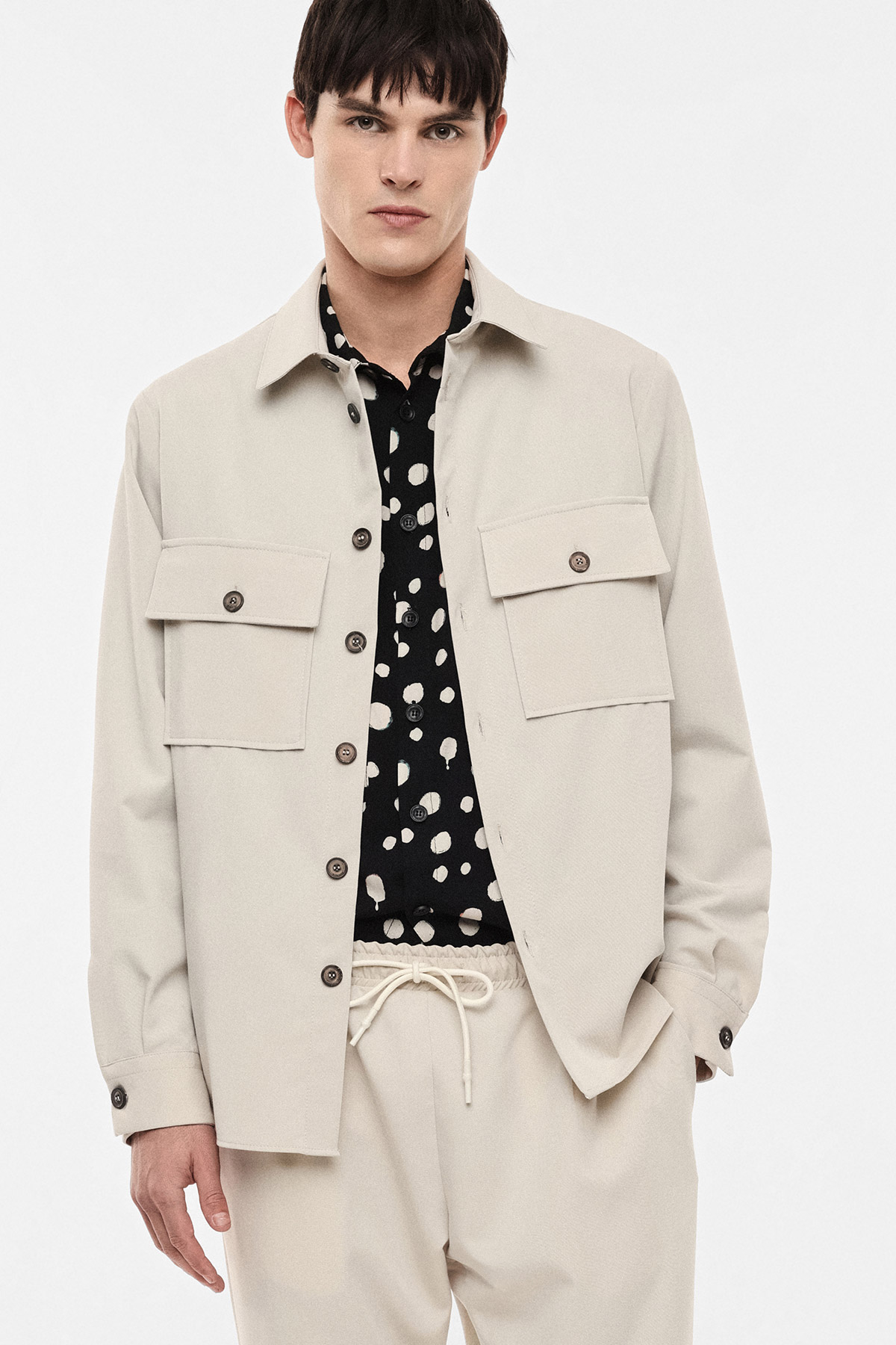 Male model wearing cream overshirt and matching trousers over a black polka-dot shirt, casual smart menswear outfit
