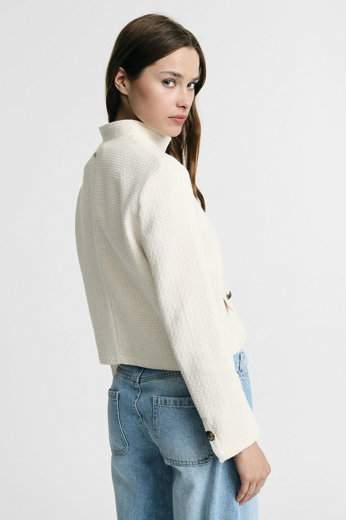 Woman looking over shoulder wearing cream textured cropped jacket and light blue jeans on plain white background