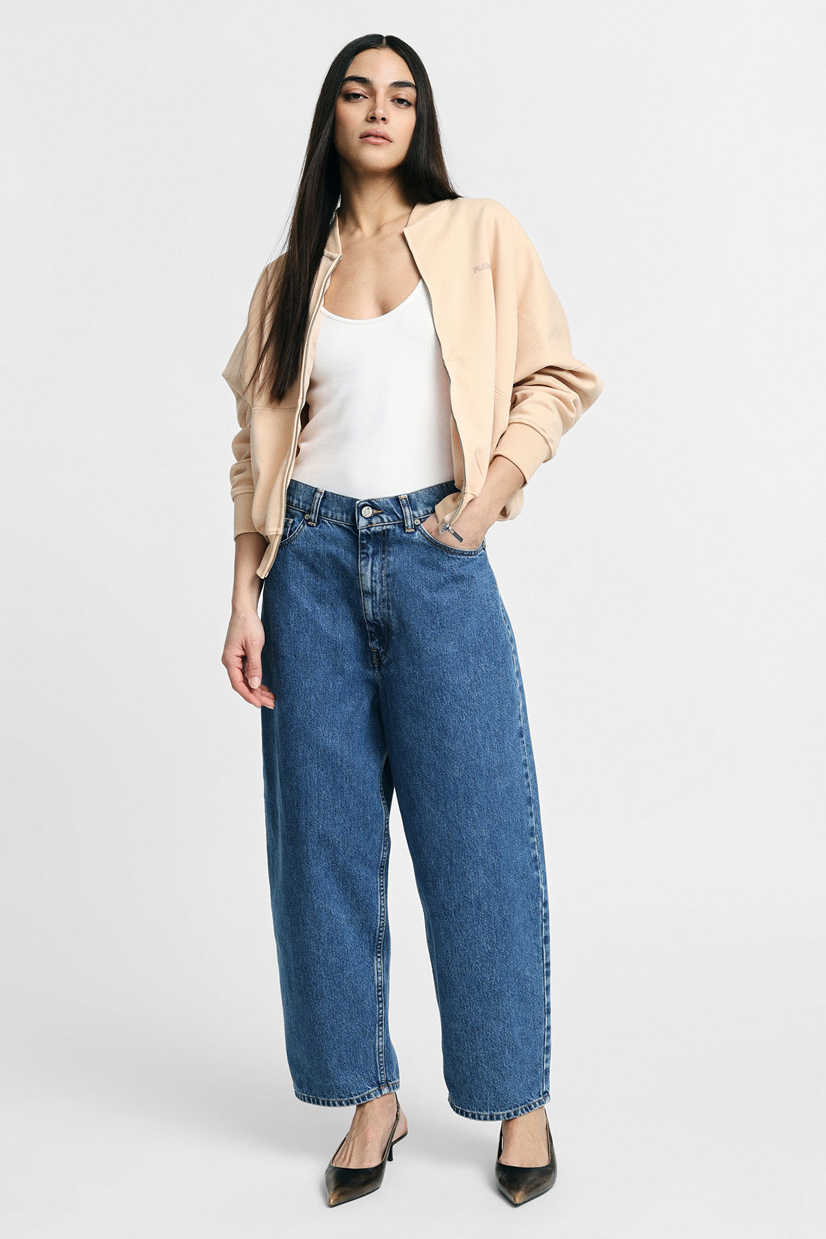 Female model standing in beige bomber jacket, white tank top, wide-leg blue jeans and black heels