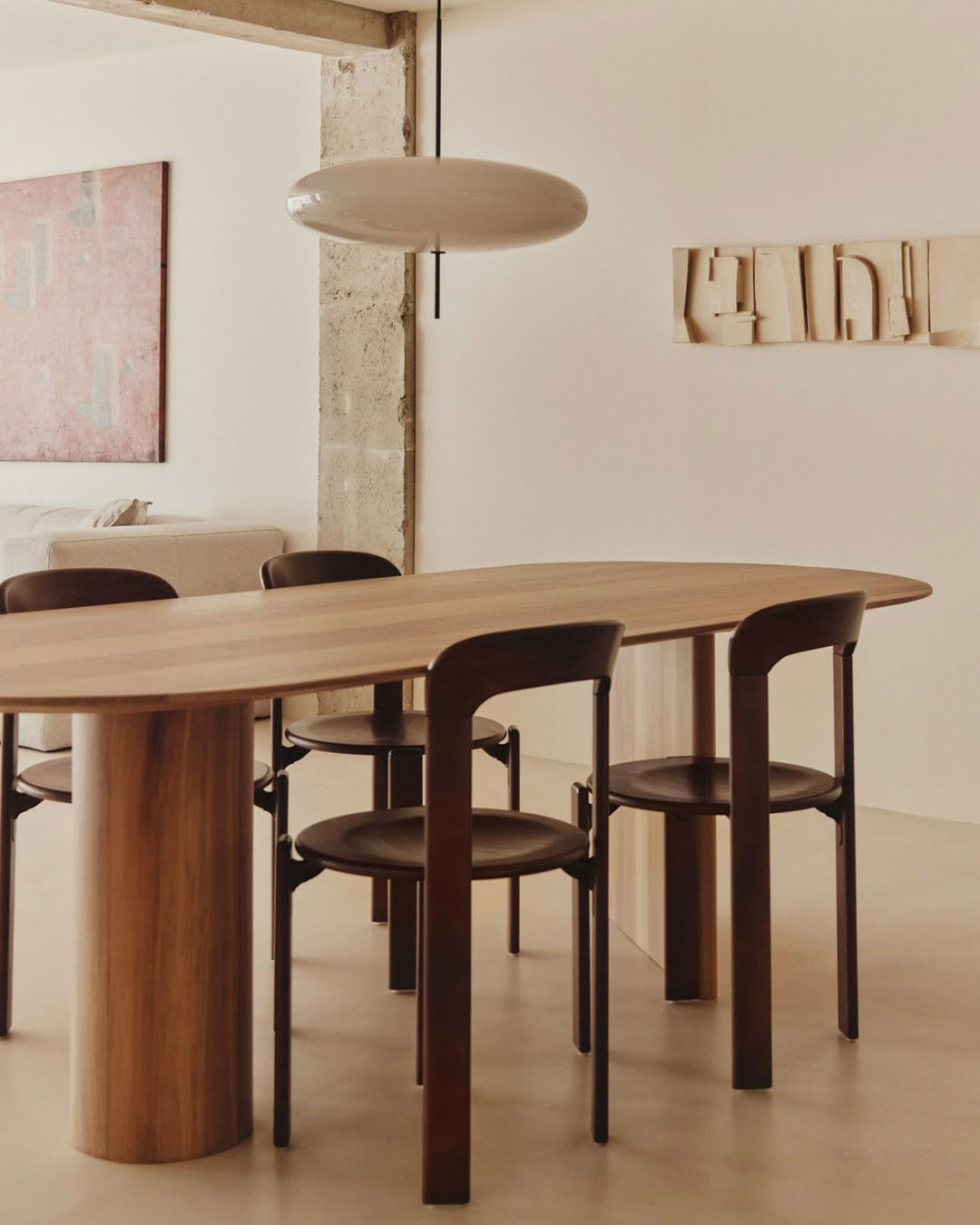 Minimalist dining room with oval wooden table, four dark chairs, pendant light, neutral walls and abstract artwork