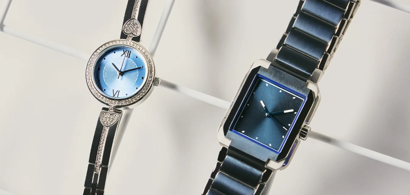 Due orologi da polso eleganti con quadranti blu e cinturini in metallo, uno rotondo con dettagli a forma di cuore, l'altro rettangolare, esposti su una superficie a griglia bianca.