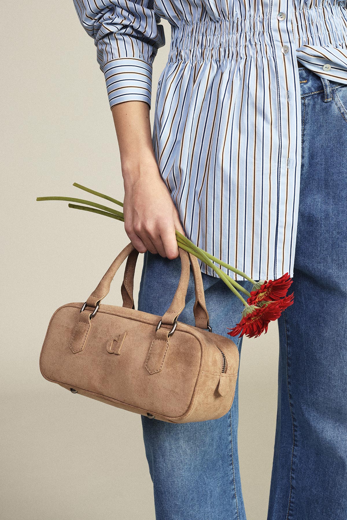 Cropped view of person in striped shirt and jeans holding tan suede handbag and red gerbera daisies