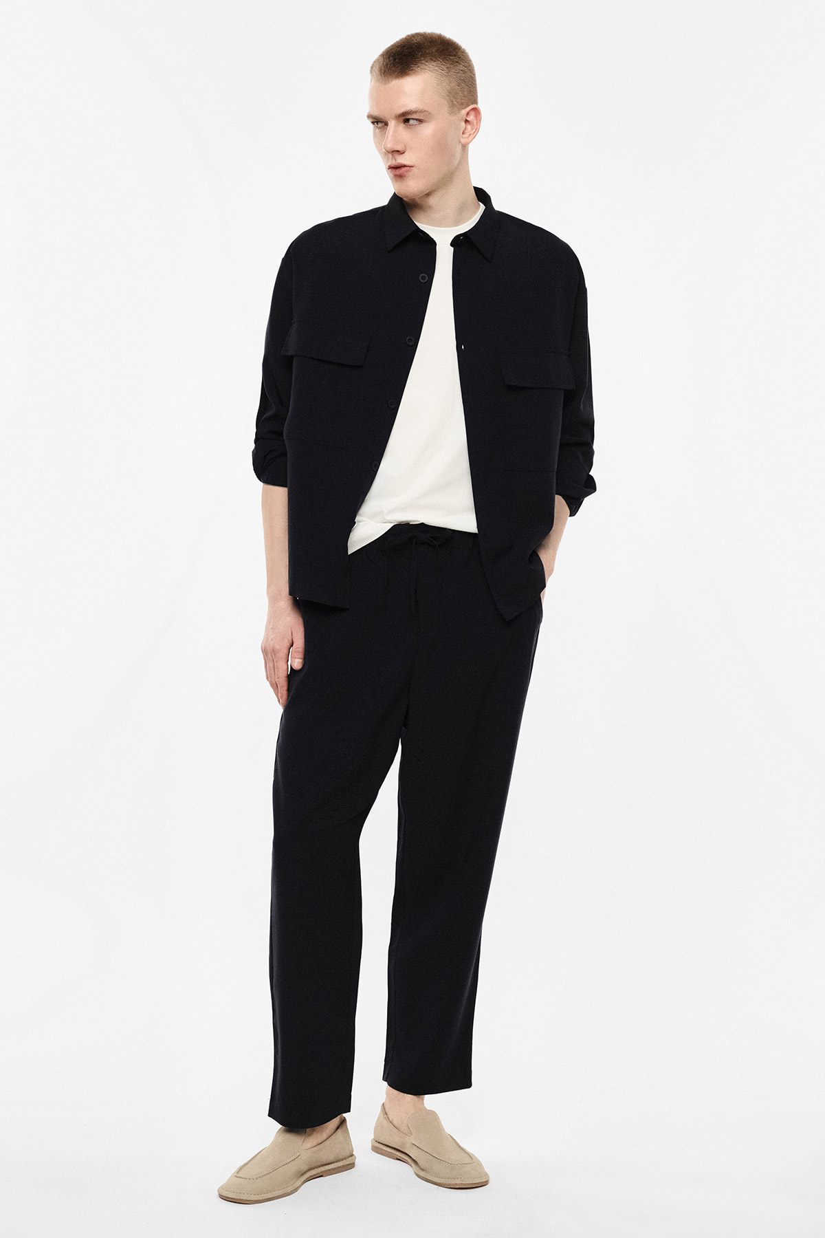 Male model in black shirt-jacket, white T-shirt, black trousers and beige loafers, standing on a white background