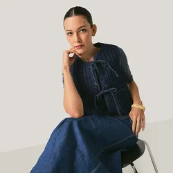 Donna seduta su una sedia, indossa un gilet di lana blu navy e una gonna lunga coordinata, poggiando il mento sulla mano, sfondo neutro.