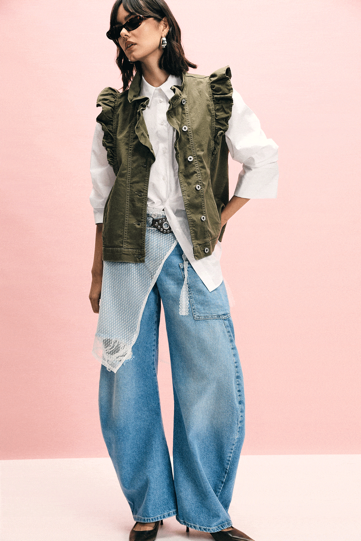 Woman wearing wide-leg distressed blue jeans with white shirt, olive ruffle vest and black heels against pink backdrop