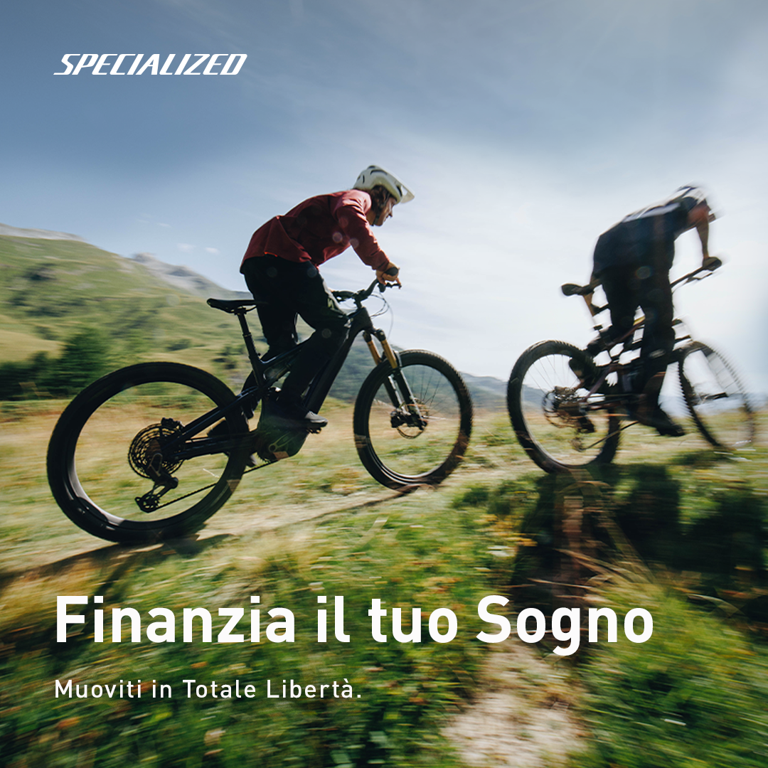 Two mountain bikers riding Specialized e-bikes on a grassy hillside trail with motion blur under a bright sky