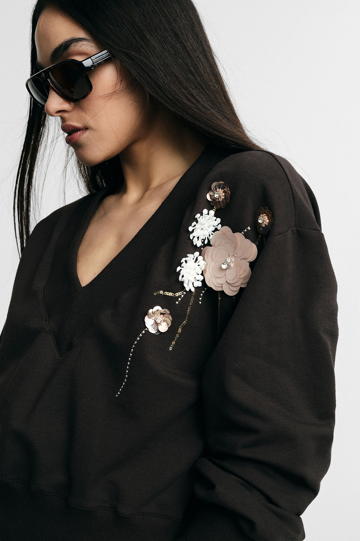 Woman in profile wearing dark brown V-neck sweatshirt with embellished floral appliqué on shoulder and sunglasses