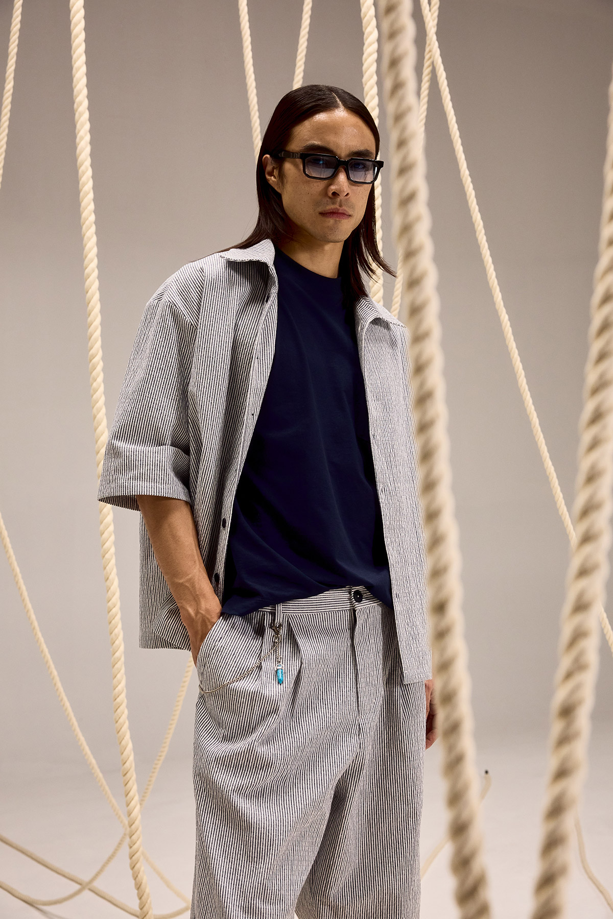 Person wearing sunglasses, navy T-shirt and matching striped shirt and trousers standing among hanging ropes in a studio