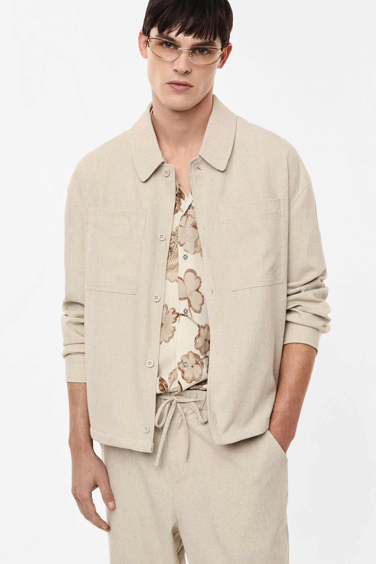 Male model wearing beige linen jacket over a floral shirt with drawstring trousers, hands in pockets on a white background