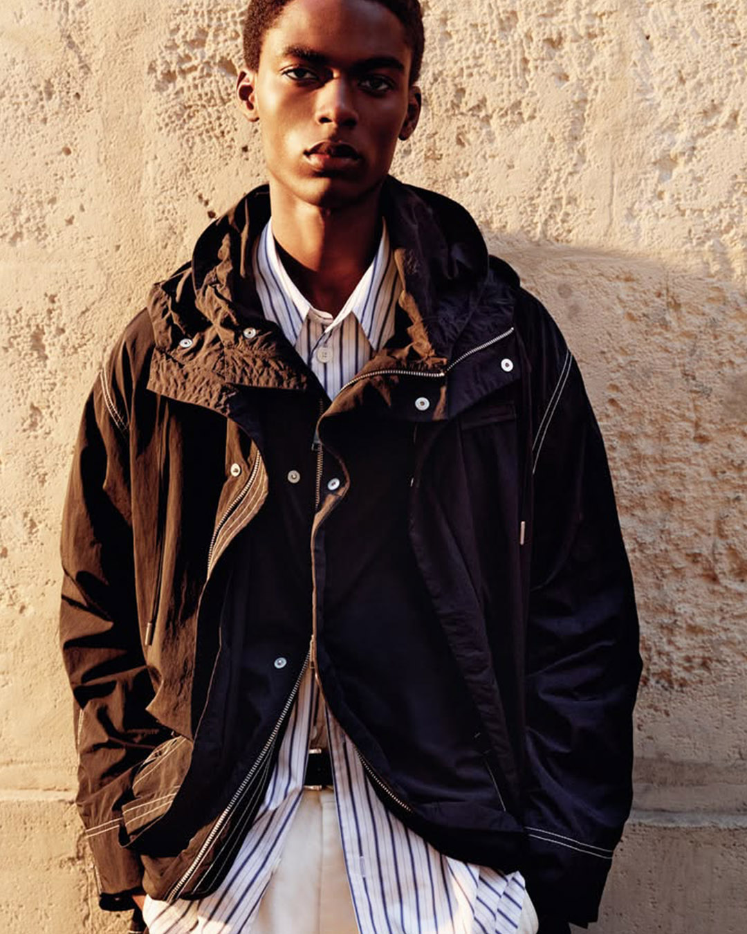 Young man wearing a black hooded jacket over a striped shirt and light trousers, standing against a textured beige wall
