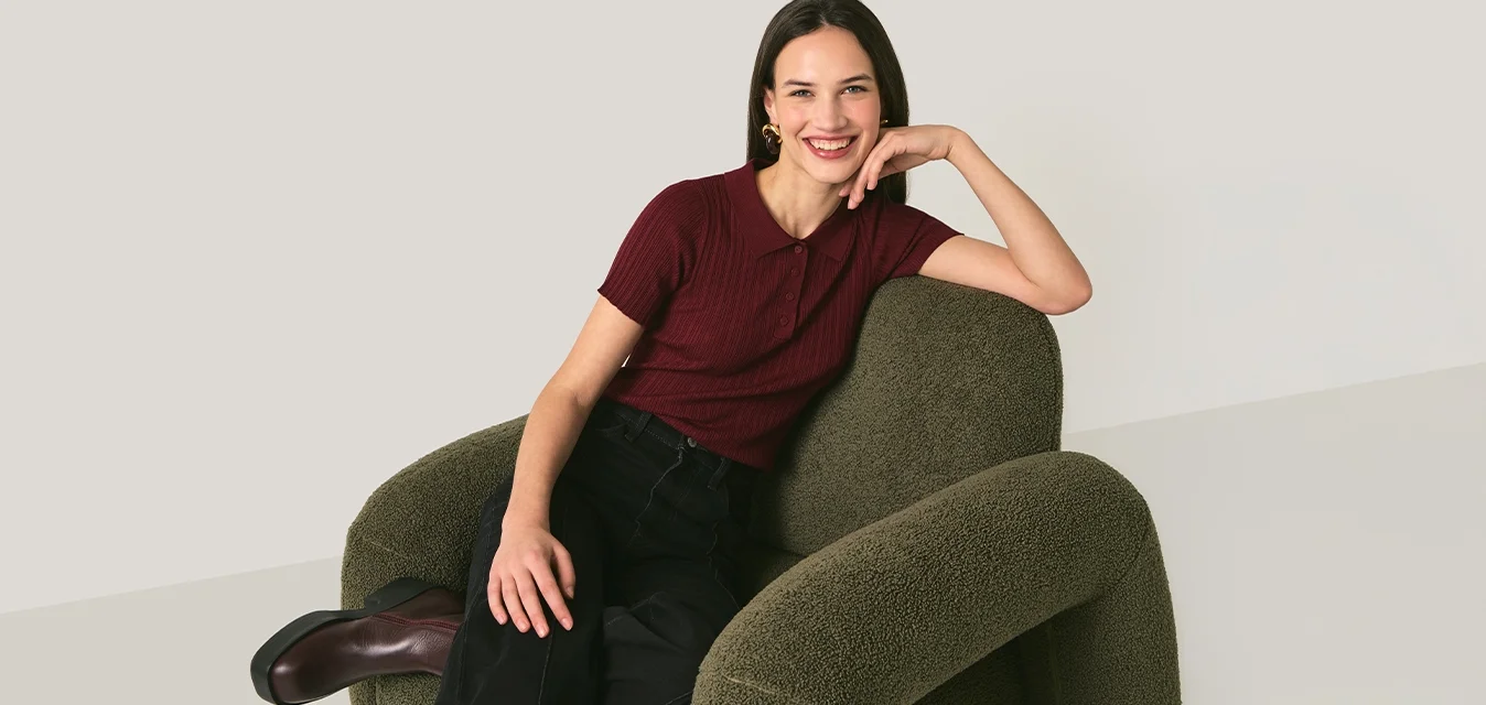 Giovane donna sorridente, seduta su una poltrona verde testurizzata, indossa una maglietta color amaranto, pantaloni neri e stivali marroni su uno sfondo semplice.