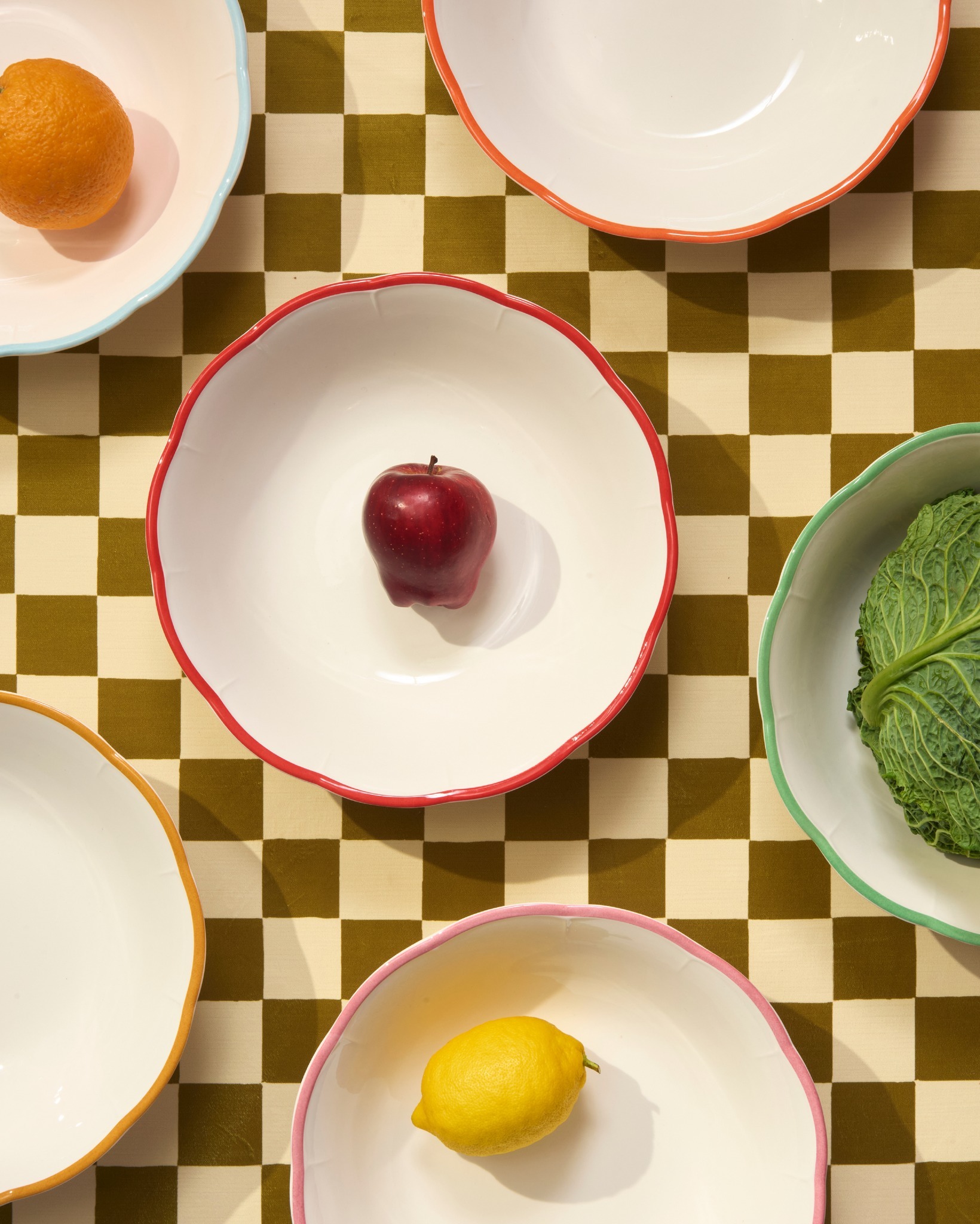 Overhead view of white scalloped bowls with coloured rims on green checkerboard cloth holding a red apple, lemon, orange and savoy cabbage