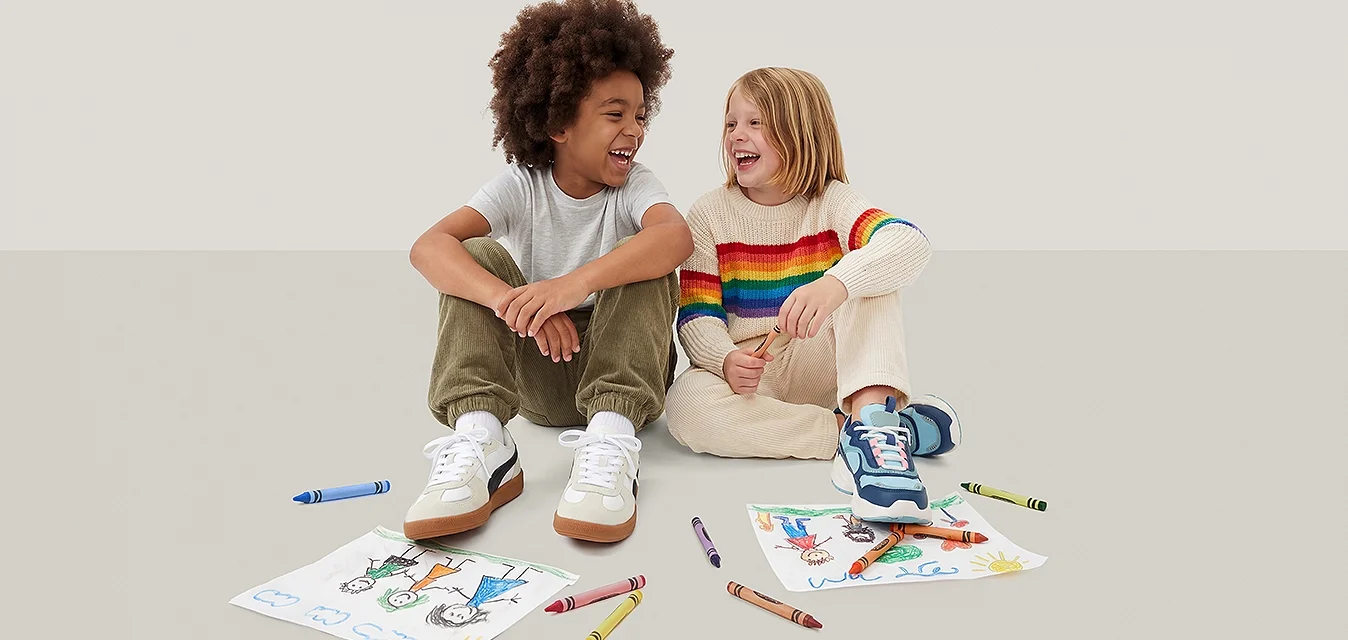 Due bambini seduti per terra ridono, circondati da pastelli colorati e disegni su carta.