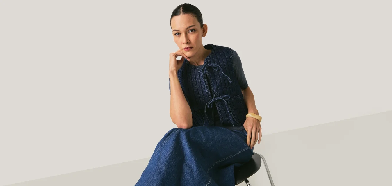 Donna seduta su una sedia, indossa un gilet di lana blu navy e una gonna lunga coordinata, poggiando il mento sulla mano, sfondo neutro.