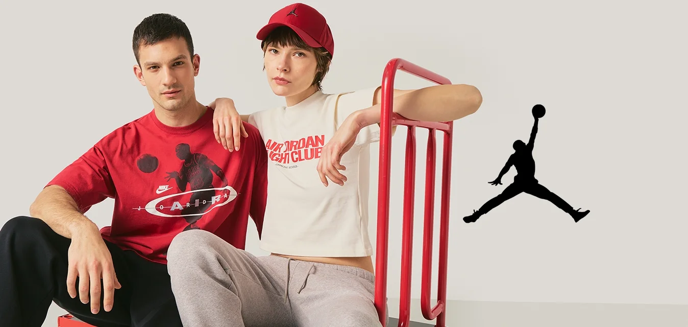 Due giovani adulti sono seduti su un carrello rosso a piattaforma; uno indossa una maglietta rossa con disegni, l'altro una maglietta bianca corta e un cappellino rosso.