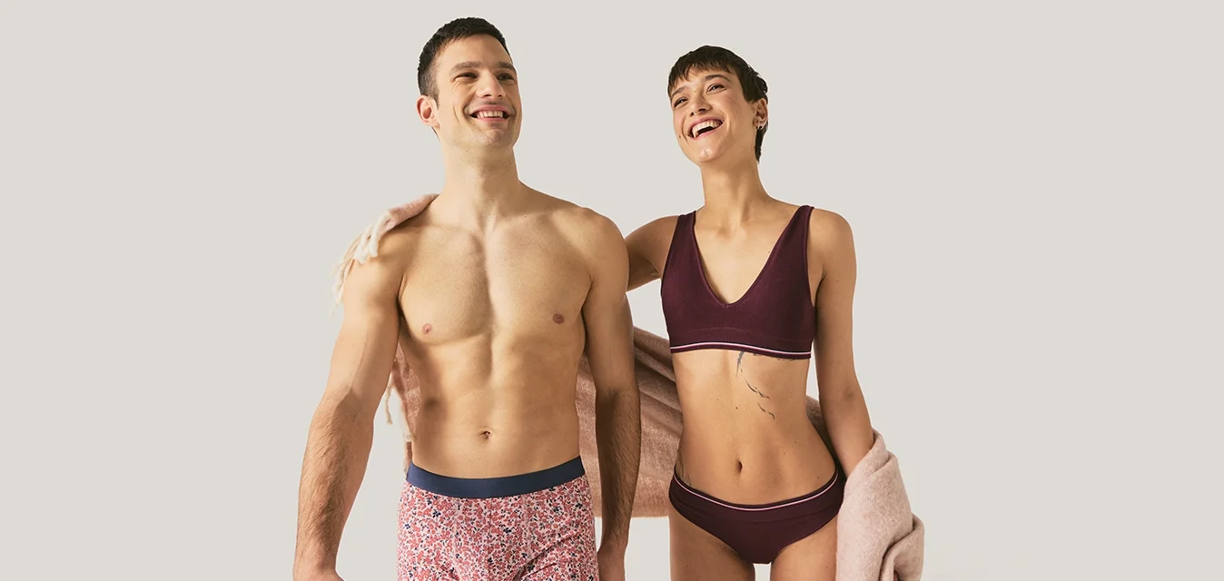 Uomo e donna sorridenti in biancheria intima, in piedi vicini; la donna avvolge una sciarpa rosa chiaro sul braccio, sfondo neutro chiaro.