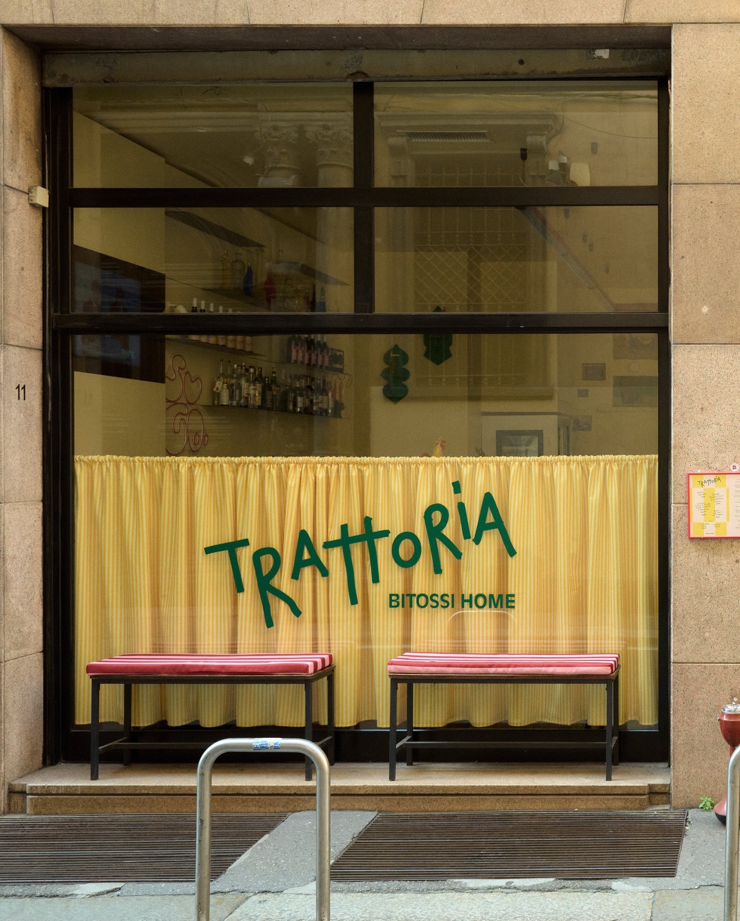 Front window of Italian trattoria 'Bitossi Home' with yellow curtain, green signage and two red-cushioned benches outside