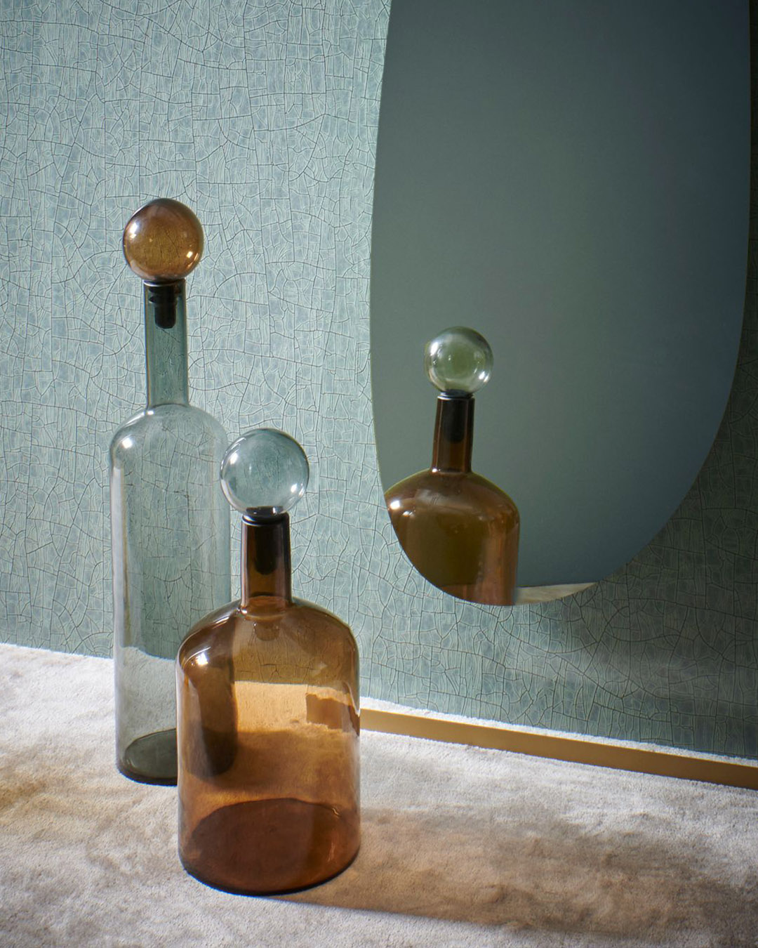 Two glass decanters with round stoppers (amber and clear) reflected in an oval mirror against teal textured wallpaper