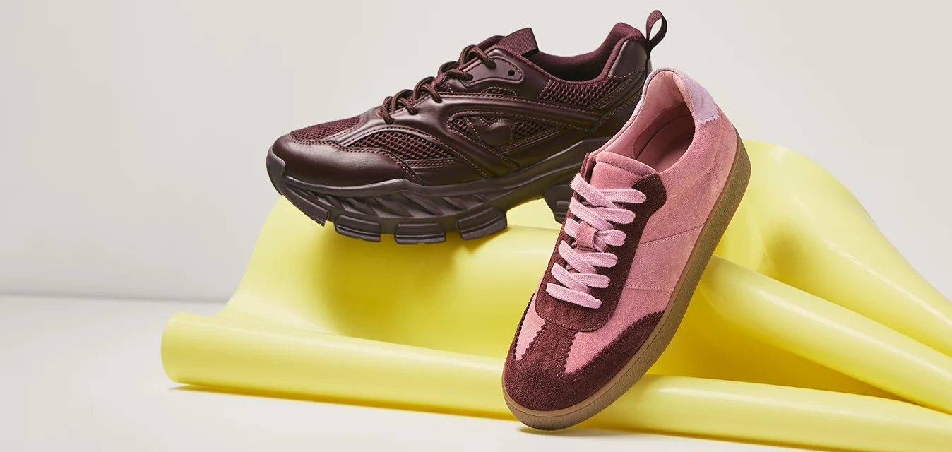 Coppia di sneakers bordeaux e rosa appoggiate su un tappetino da ginnastica giallo piegato su uno sfondo semplice.