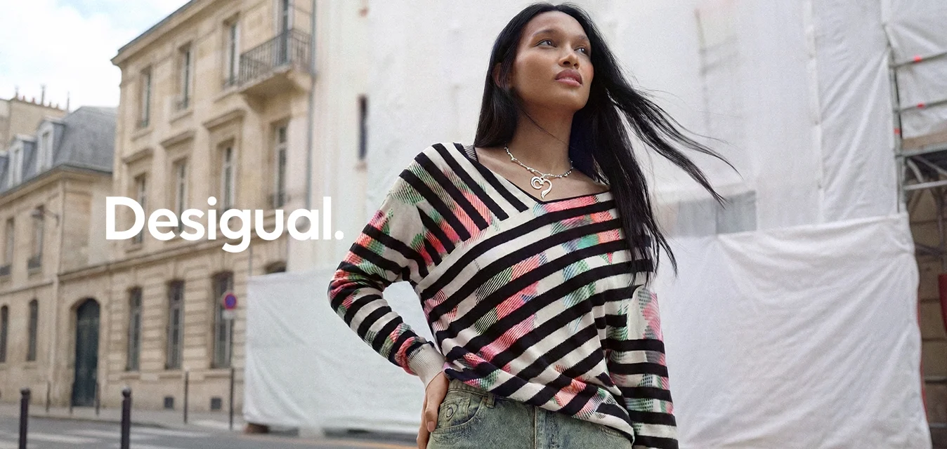 Donna con capelli lunghi e scuri che indossa un maglione a righe multicolore e una gonna di jeans, in piedi in una strada della città con edifici storici.