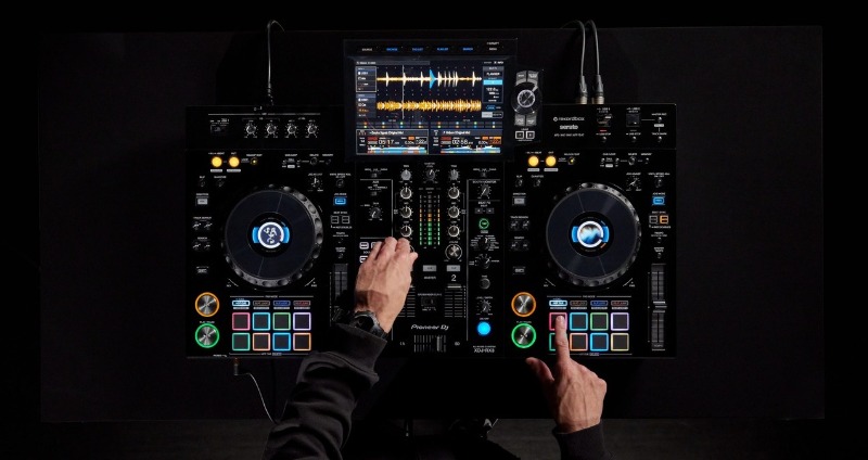 Top-down view of a DJ controller with colourful pads and waveform display, hands adjusting knobs and cueing tracks