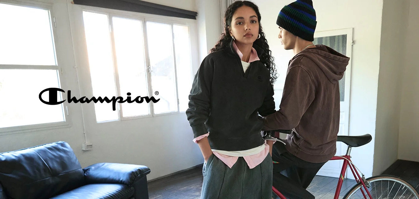 Due persone al chiuso accanto a una bicicletta rossa; una è in piedi con le mani in tasca, l'altra è seduta sulla bici con un berretto a righe in una stanza spaziosa.