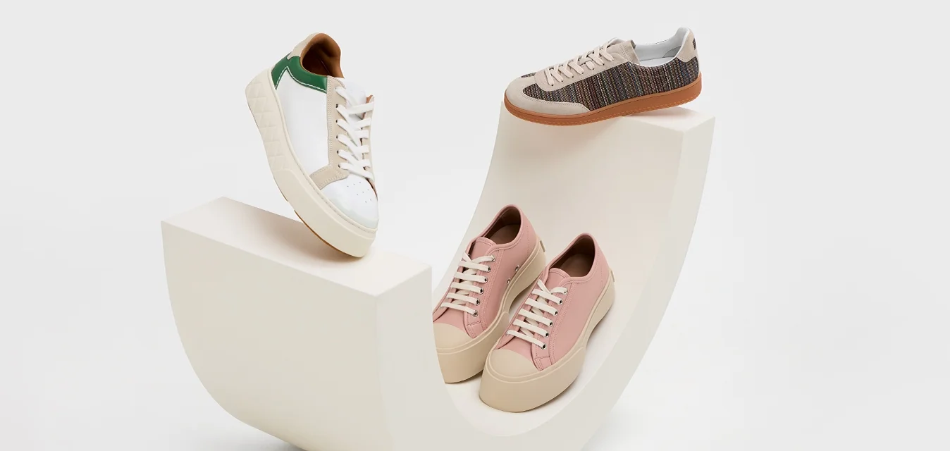 Sneaker casual bianchi con accenti verdi, rosa chiaro e texture intrecciate marroni, esposte su una piattaforma curva bianca.