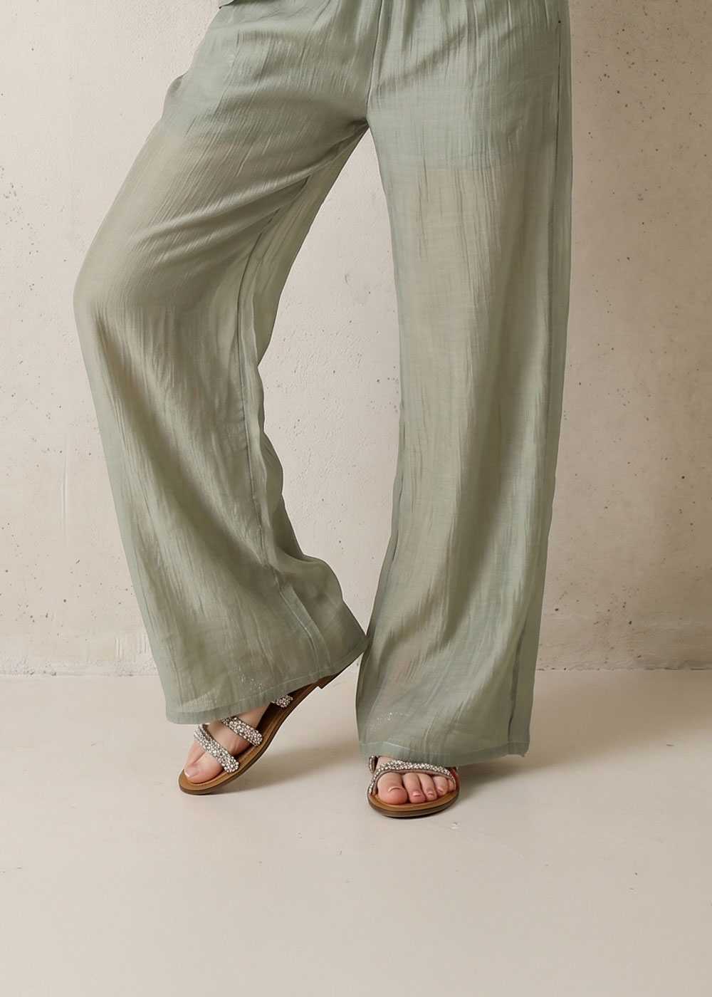 Model wearing sage-green wide-leg linen trousers and beaded flat sandals on a neutral studio background