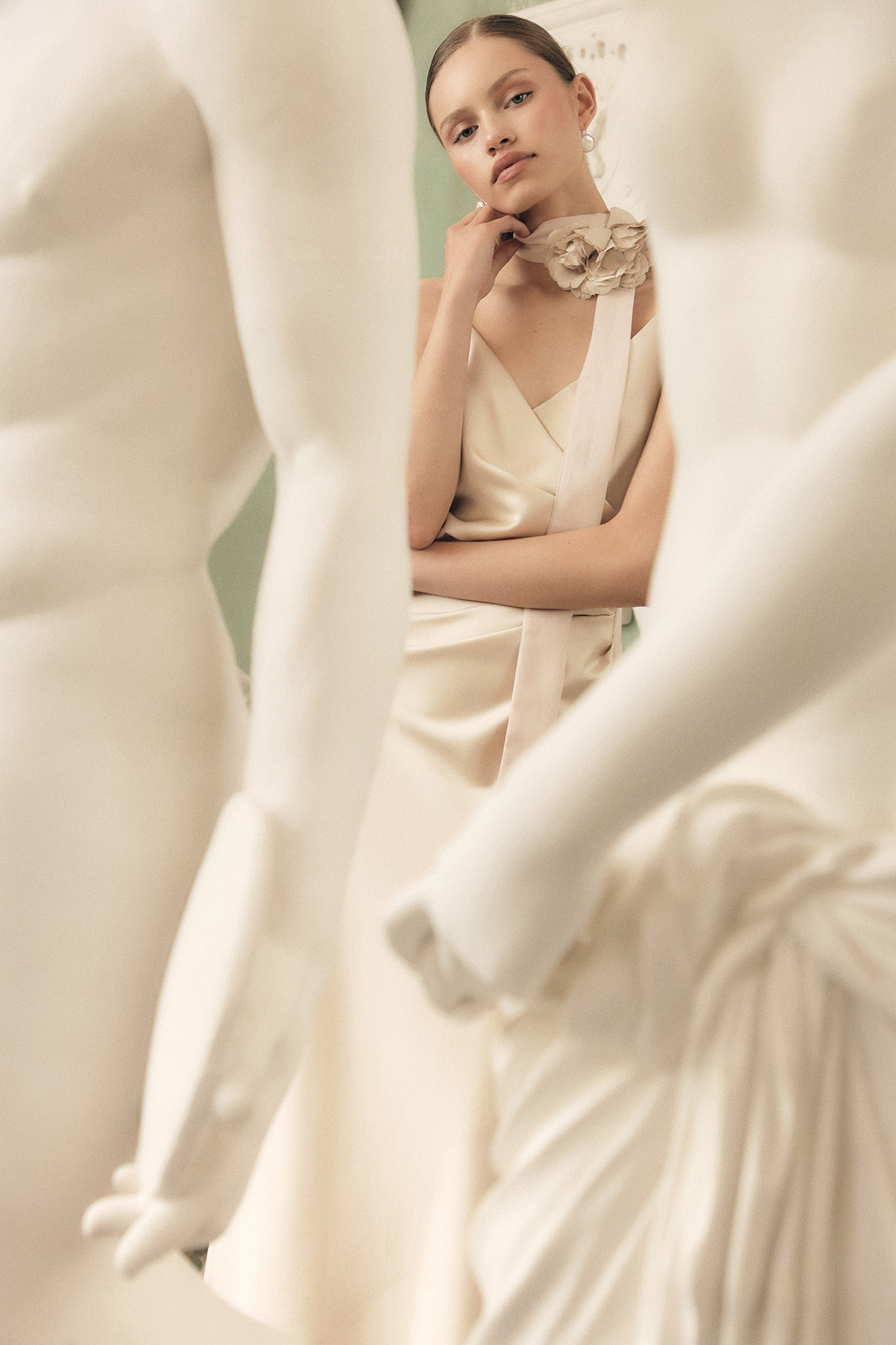 Young woman in cream satin dress with floral neckpiece posed among white mannequins in a fashion editorial
