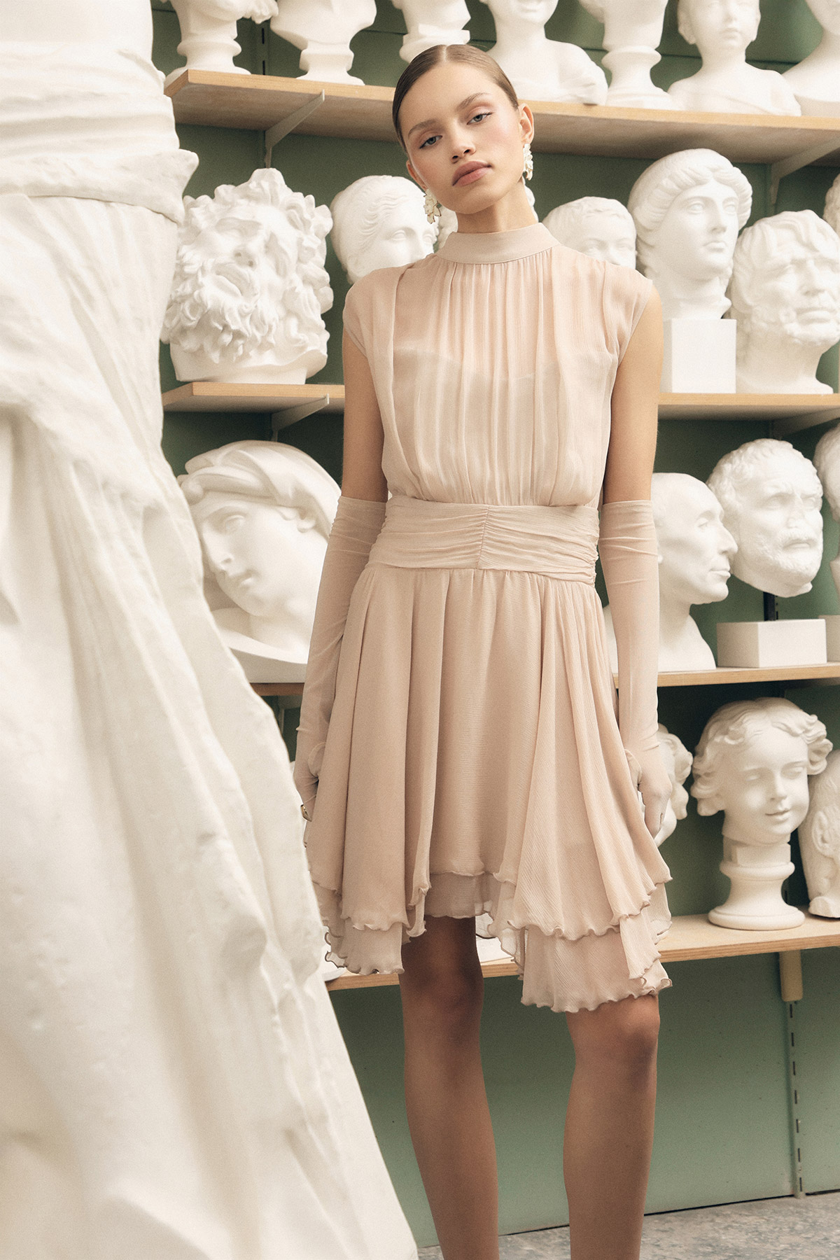 Model in a pale blush chiffon dress and long gloves standing in a sculptor's studio surrounded by plaster busts