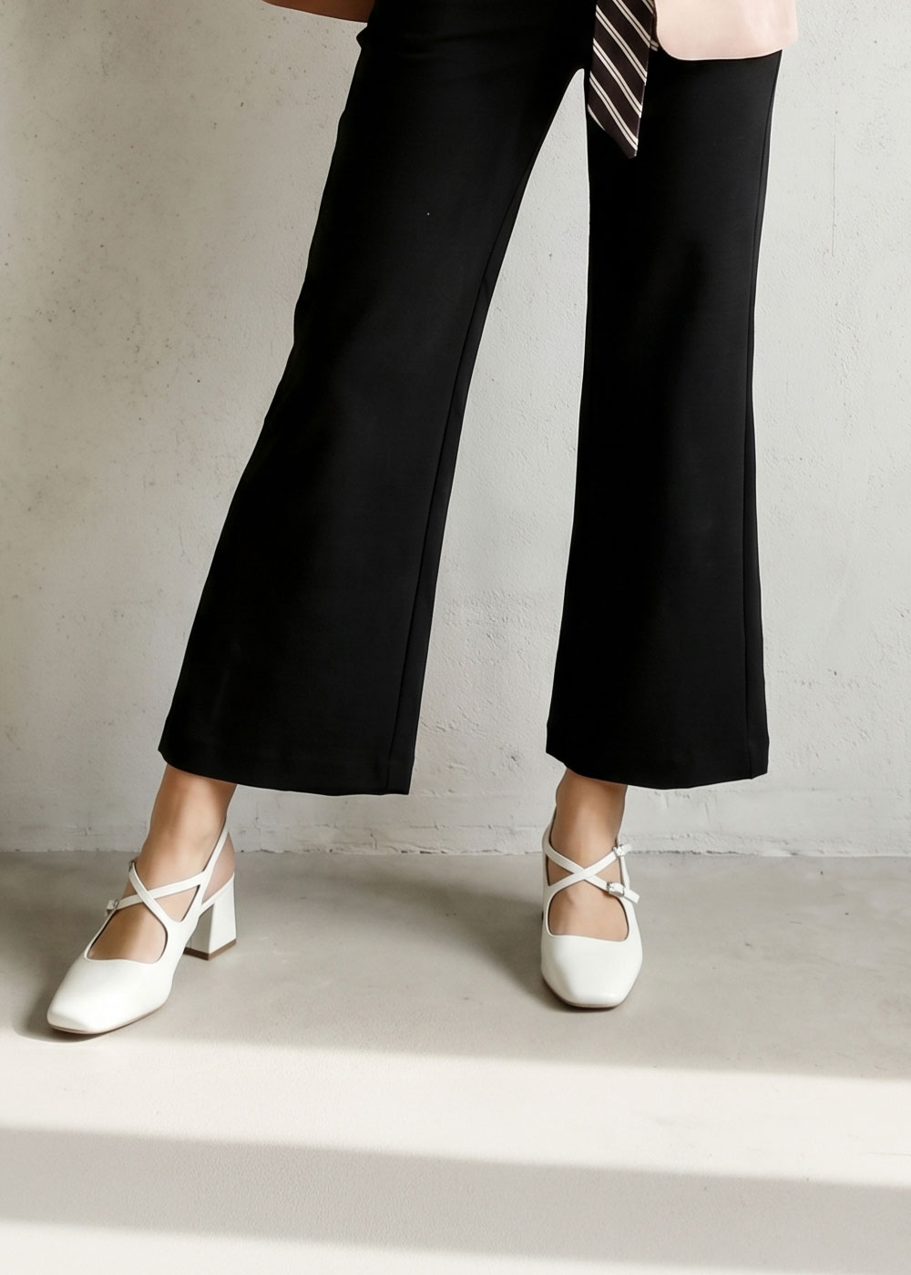 Cropped view of legs in black wide-leg trousers wearing white strappy block-heel shoes against a pale wall