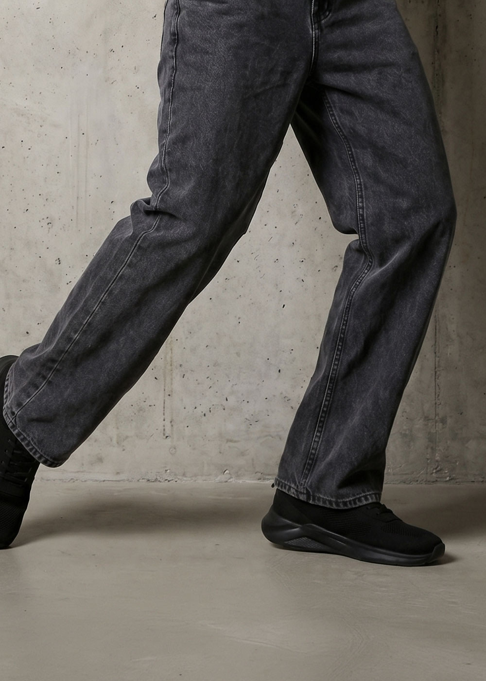 Person wearing faded black wide‑leg jeans and black trainers stepping on a concrete floor against a raw concrete wall
