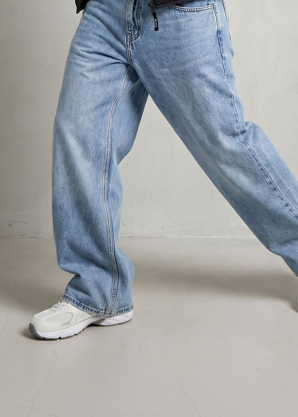 Person wearing light-blue wide-leg denim jeans and white trainers stepping forward on a plain studio floor, cropped view of legs