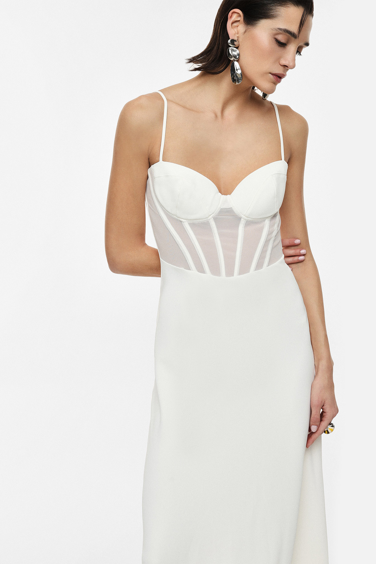 Model wearing a white fitted slip dress with sheer corset bodice and thin straps, statement earrings