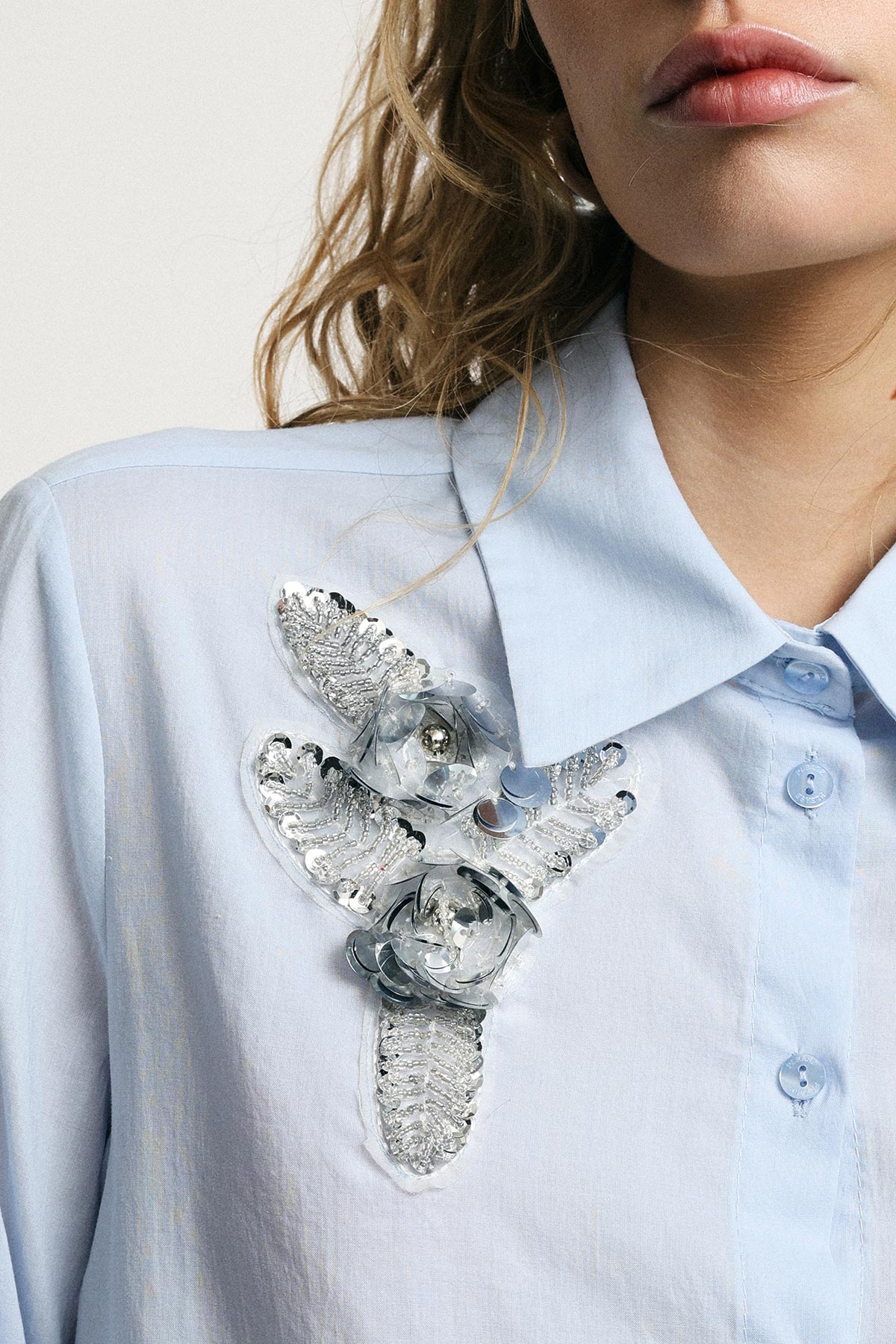 Close-up of woman wearing a pale blue collared shirt with silver sequin floral appliqué on the chest, showing lips and hair