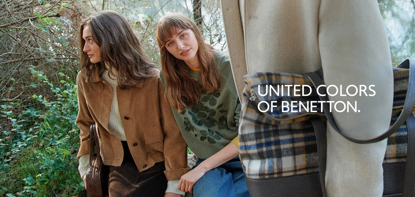 Tre persone vicino al lago, due sedute su una panchina con giacche casual, una in piedi con un cappotto beige che tiene una borsa a quadri.