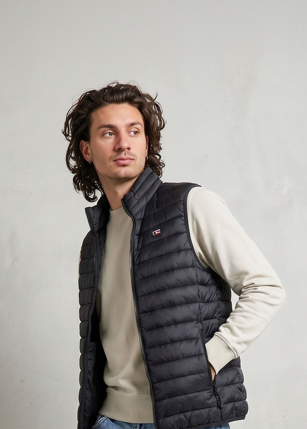 Male model wearing a black quilted puffer gilet over a beige sweatshirt, hands in pockets, looking left against a light grey background