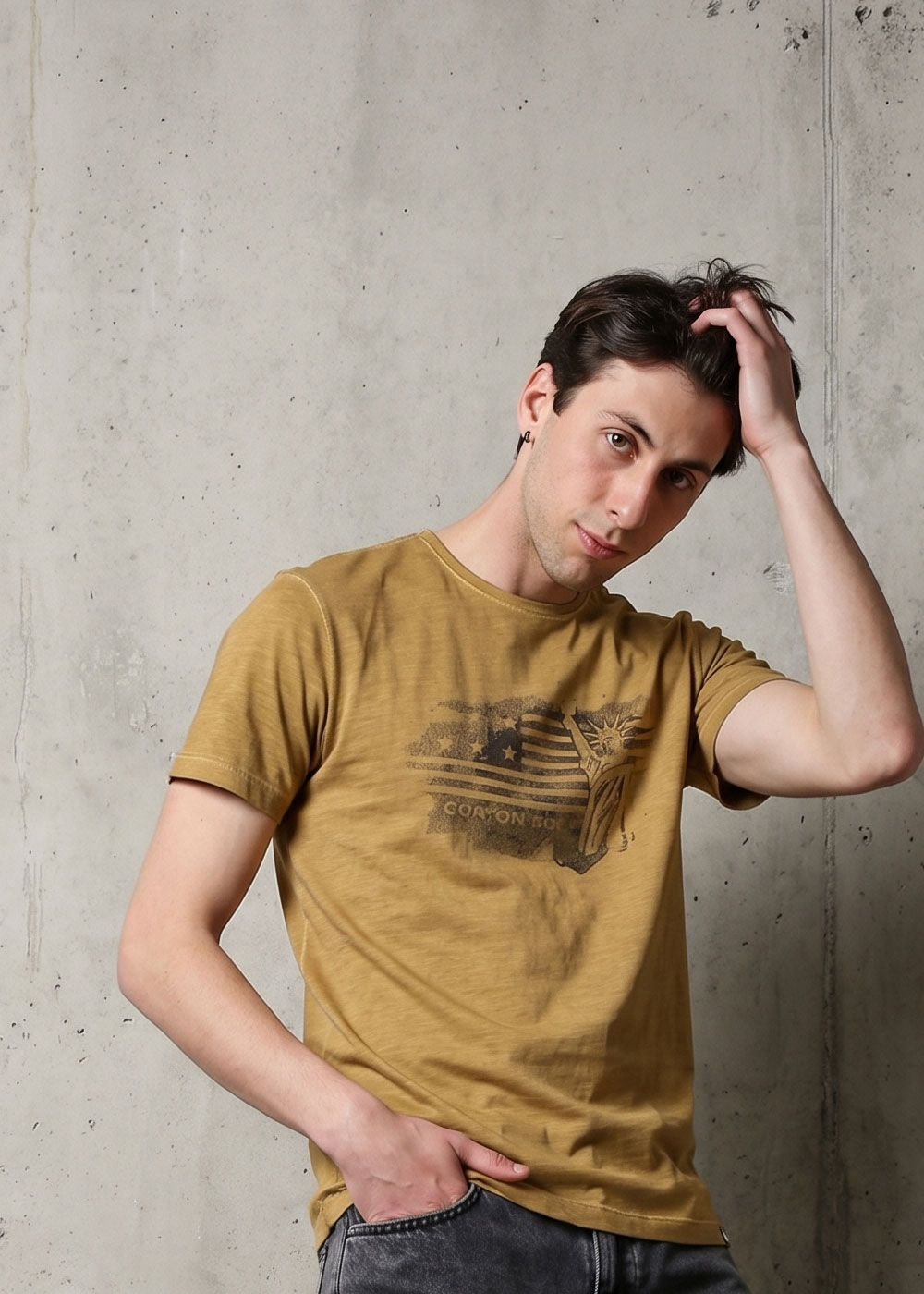 Young man in mustard t-shirt and jeans posing against a concrete wall, hand in hair, casual male model photo