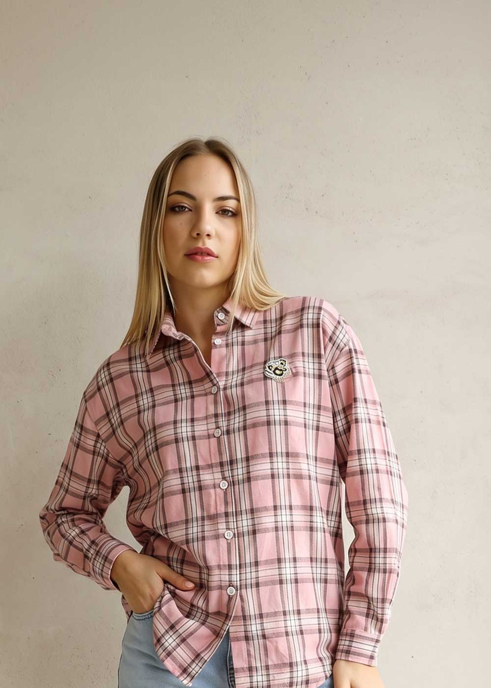 Young woman wearing a pink plaid button-up shirt and light jeans standing against a pale textured wall