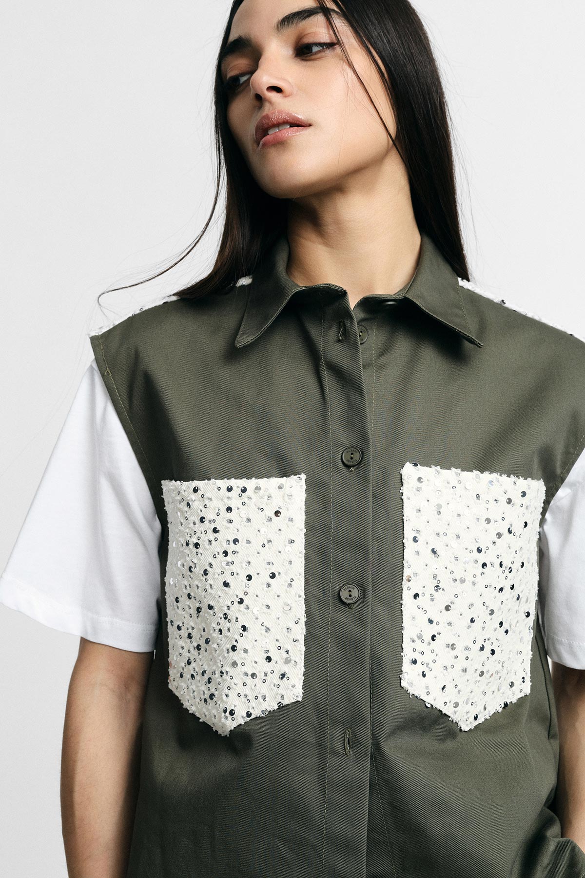 Young woman modelling sleeveless olive-green button-up vest with white sequin patch pockets over a white T‑shirt