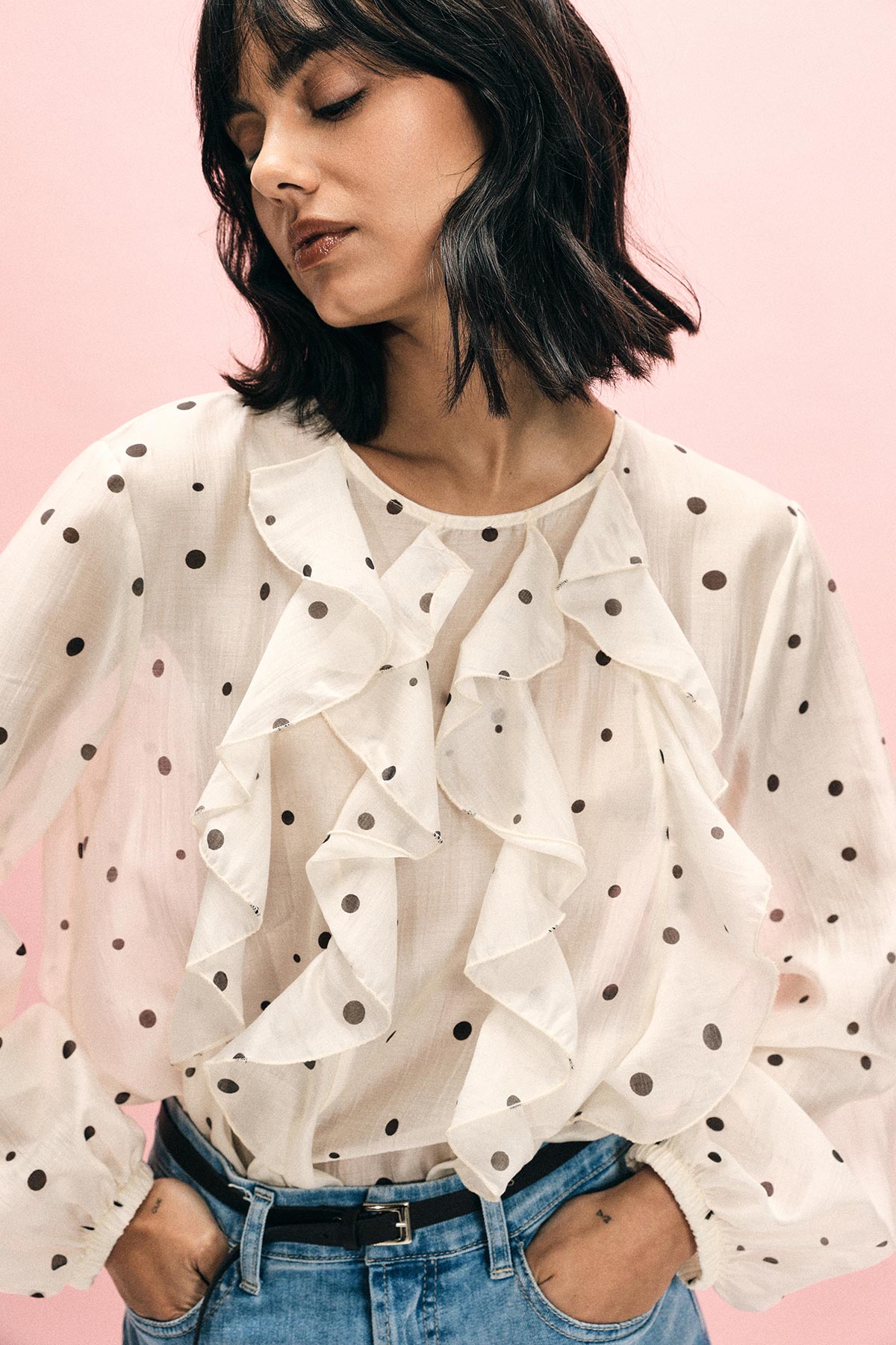 Woman wearing sheer white polka-dot ruffled blouse tucked into jeans, hands in pockets against a pink background