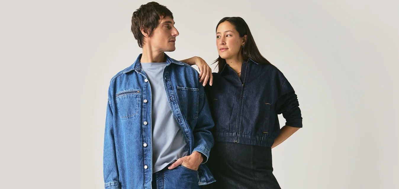 Uomo e donna in piedi uno di fronte all'altro, entrambi vestiti con abiti in denim scuro e scarpe nere, su uno sfondo grigio chiaro uniforme.